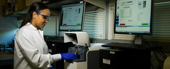 Scientist in lab coat using specialized equipment with data displayed on computer monitors.