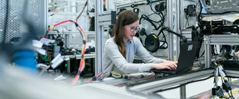 Ingenieur arbeitet an Laptop, umgeben von Maschinen und Kabeln in einem Industrie-Labor.